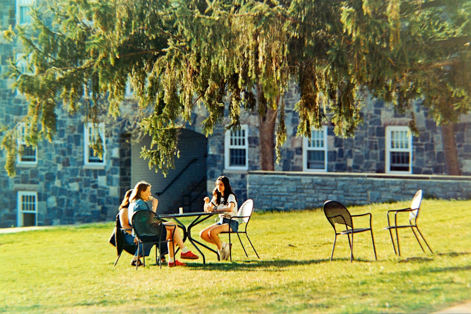 Two students talk over books at a small table on a campus lawn