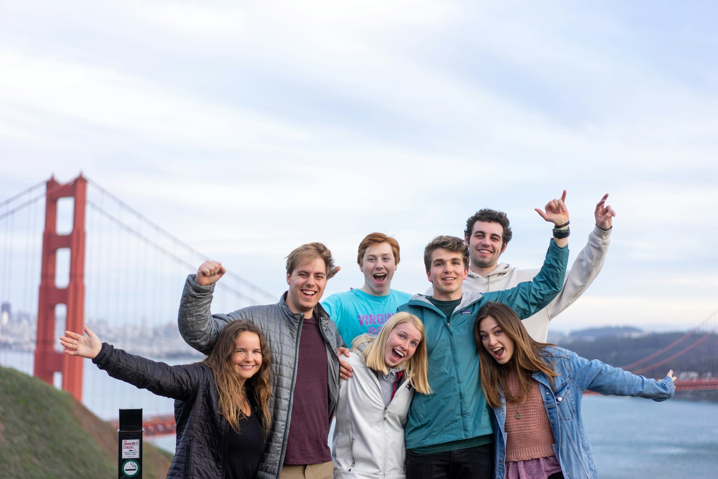 A group of seven smiling friends cheer with arms raised in front of the Golden Gate Bridge on a cloudy day