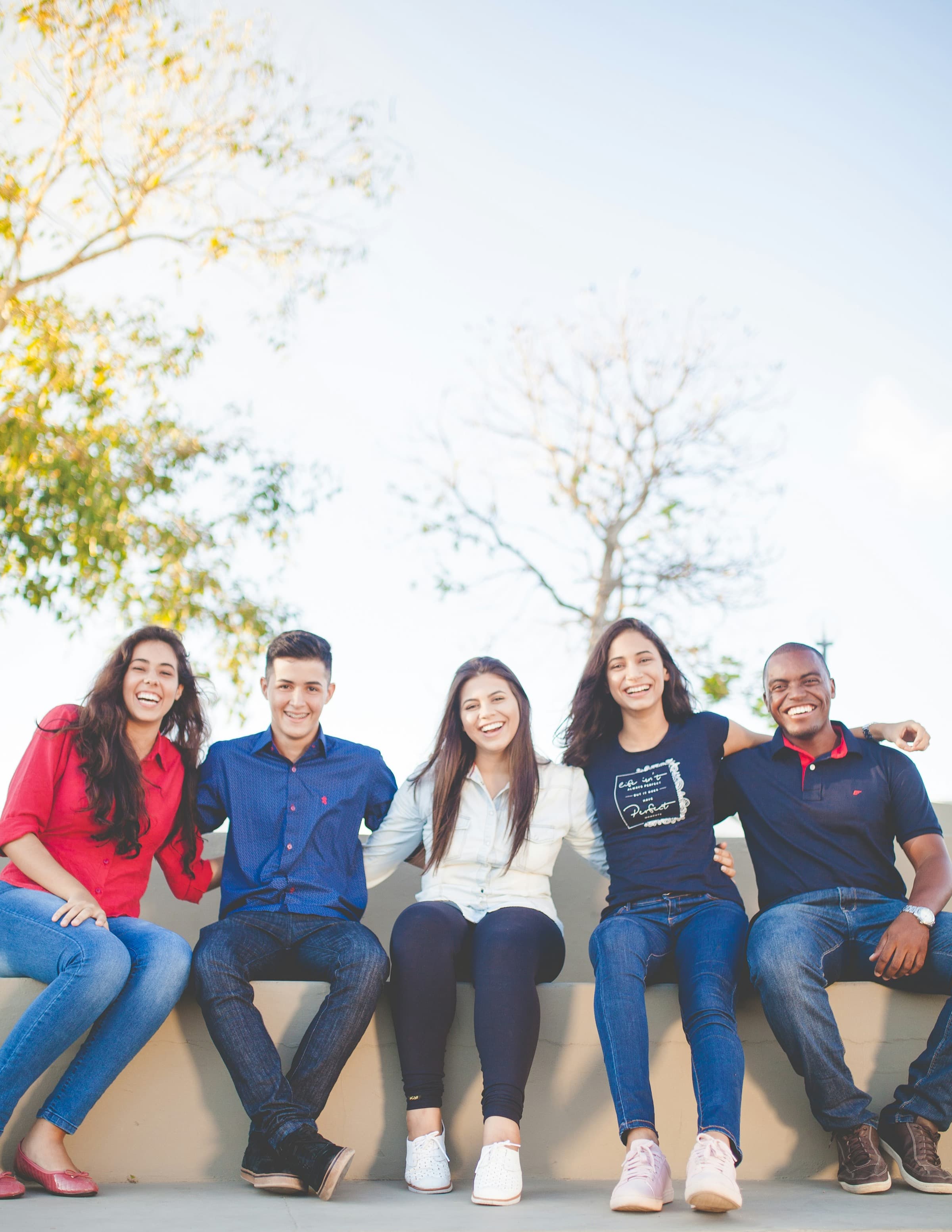 Five smiling students sit side by side on a low wall with their arms around each other, laughing at the camera against a bright sky